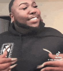 a man with a beard and long nails is wearing a black hoodie and smiling .