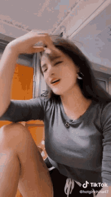 a woman in a gray shirt is sitting on a table with her legs crossed and making a funny face .