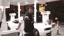 a man is pointing at the camera while standing in front of a gas station .