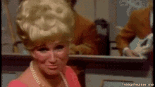 a woman with blonde hair and a pearl necklace is looking at herself in the mirror .