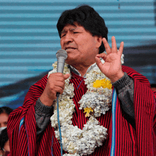 a man with flowers around his neck is speaking into a microphone with his hand up