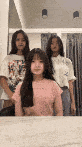 three girls are standing in front of a mirror and one of them is wearing a pink shirt