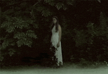 a woman in a white dress is standing in front of a forest .
