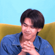 a young man in a denim shirt is sitting on a yellow chair with his hands folded