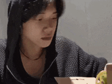 a man wearing a hoodie is sitting at a table looking at a laptop .