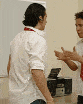 a man in a white shirt and red tie talks to another man in front of a stereo