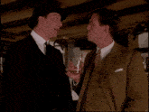 a man in a suit is holding a glass and talking to another man in a hat
