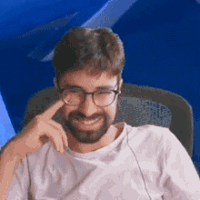 a man with a beard and glasses is sitting in a chair with his hand on his chin .