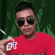 a man wearing sunglasses and a red shirt is smoking a cigarette and has a #t sticker on his shirt .