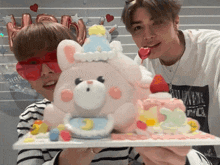 two young men holding a cake with a teddy bear on top