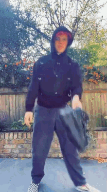 a person wearing a black hoodie and black pants is standing in front of a wooden fence holding a bag .