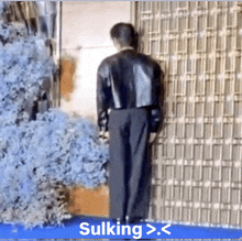 a man in a black leather jacket is standing in front of a building with the words sulking > . < on the bottom right