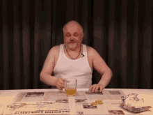 a man in a white tank top is sitting at a table with a glass of beer and a bag of chips .