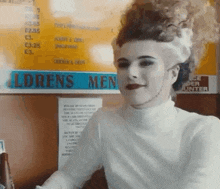 a woman in a white turtleneck is sitting in front of a sign that says ldrens men .