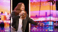 a man with long hair singing into a microphone with a nbc logo in the corner