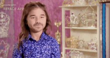 a man with long hair and a beard wearing a blue shirt is standing in front of a shelf .