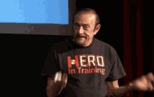 a man with a mustache wears a black shirt that says hero in training