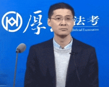 a man in a suit stands in front of a microphone in front of a blue wall that says judicial examination