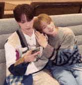 two young men are sitting on a couch one is holding a cell phone and the other is hugging him