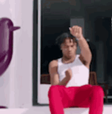 a man in a white tank top and red pants is sitting on a chair with his fist in the air .