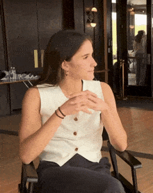 a woman in a white vest sits in a chair with her hands folded
