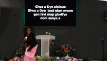 a woman stands in front of a podium with the words gwa a dye aleloua written on the screen behind her