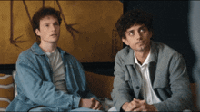 two men are sitting on a couch and one of them is wearing a grey jacket