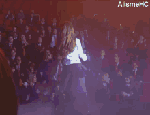 a woman in a skirt stands in front of a crowd of people with the alismehc logo in the corner