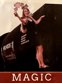 a woman in a black dress is holding a bunch of playing cards in front of a sign that says " magic "