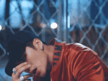 a man wearing a red jacket and a black hat is sitting in front of a chain link fence with his hand on his face .