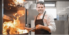 a man in an apron is cooking food in a kitchen with flames behind him