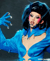 a drag queen is wearing a blue top and gloves and has a necklace around her neck