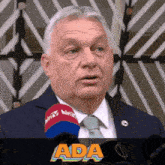 a man wearing a suit and tie is speaking into a microphone with the word ada behind him