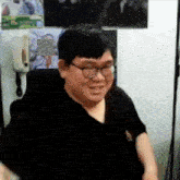 a man wearing glasses and a black shirt is sitting in front of a wall with pictures on it