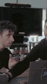 two men are sitting at a table with a laptop and a phone with a sticker that says sc on it