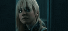 a woman with blonde hair and a collar around her neck looks at the camera in a dark room .
