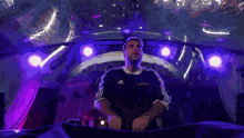 a man wearing an adidas shirt stands in a dark room with purple lights