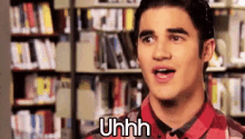 a man in a red plaid shirt is standing in front of a bookshelf in a library and saying uhh .