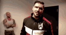 a man in a black and red jacket is standing next to another man with his arms crossed in a room .