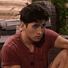 a young man in a red t-shirt is sitting on a bench and looking at the camera .