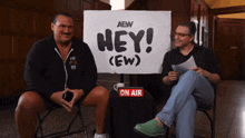 two men sitting in front of a sign that says hey