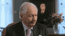an older man in a suit and tie is sitting in front of a painting of mona lisa