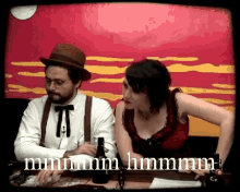 a man and a woman are sitting at a table with the words mmmm hmm written on the screen behind them