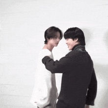 a man in a white sweater is hugging another man in a black sweater in front of a white wall