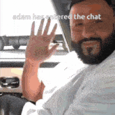 a man with a beard is sitting in a car and waving his hand with the caption adam has entered the chat