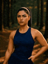 a woman in a blue tank top stands in the woods with her hands in her pockets