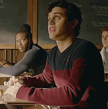 a man in a red and black sweater is sitting at a desk in a classroom with other students .