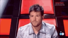 a man in a denim jacket is sitting in a red chair on a tv show .