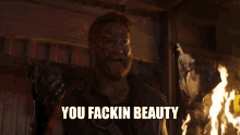 a man with blood on his face says " you fackin beauty " in front of a fire