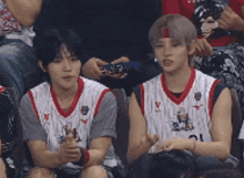 two young men in basketball uniforms are sitting in the stands watching a game .